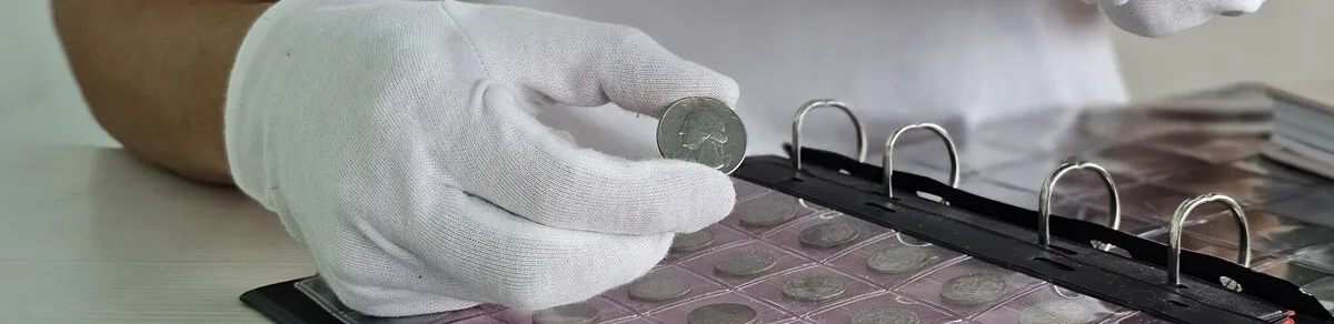 Gloved collector holding a silver coin above an album tray of stored coins